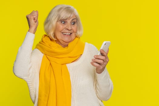 Happy excited senior woman grandmother use smartphone typing browsing shouting say wow yes found out great big win good news lottery goal achievement celebrating success, winning play game on yellow