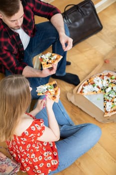 The girl sits on the floor with her legs crossed. She is wearing a red blouse and tight jeans. Next to her sits a boy with a beard and mustache. Both are holding a slice of pizza each.