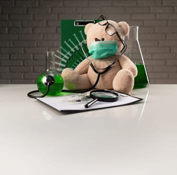 a teddy bear in a medical mask with syringes in his shoulder on the white table of a vaccine researcher.