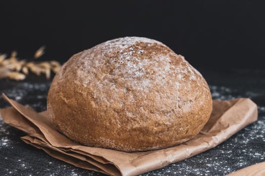 One round bread with cellulose on a paper bed and ears lies on a black stone table sprinkled with flour, side view, close-up. The concept of baking bread.
