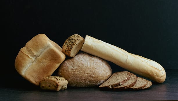 Two breads of different varieties, shapes, one half of a French baguette, buns and cut pieces lie in the center on a black stone table, close-up side view. The concept of baking bread.