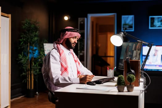 Arab entrepreneur participates in online video sessions at the office table. While using a digital desktop pc for communication and research a Muslim man pays great attention to his coworkers.