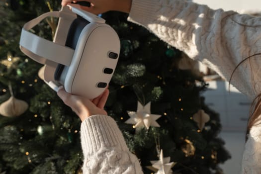 In front of a Christmas tree, a girl is holding a virtual reality headset. High quality photo