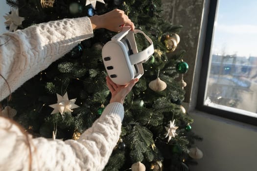 Against the setting of a Christmas tree, a girl holds a virtual reality headset in her hands. High quality photo