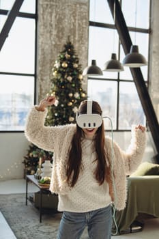 Against the backdrop of a Christmas tree, a beautiful young girl dons a VR headset. High quality photo