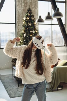 In front of a Christmas tree, a lovely young lady wears a virtual reality headset. High quality photo