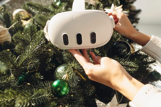 Against the setting of a Christmas tree, a girl holds a virtual reality headset in her hands. High quality photo