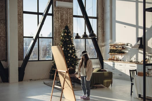 In a festive Christmas ambiance, a young lady in light clothing and a virtual reality headset gazes at herself in the mirror. High quality photo