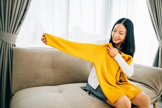 Smiling Asian woman unwraps a shirt from a box excited at home. Delighted customer reveals online package exhibiting new clothes. Delivery and online shopping concept.