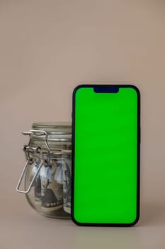 Vertical Green screen on modern mobile phone in background of glass jar full of American currency dollar banknotes on beige background. Cope space for text. Advertisement for application website. Concept of money economy banks and finances
