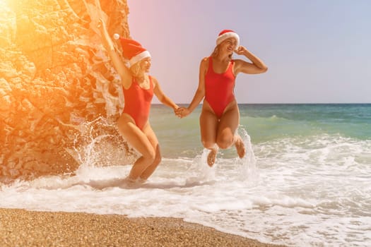 Women Santa hats ocean play. Seaside, beach daytime, enjoying beach fun. Two women in red swimsuits and Santa hats are enjoying themselves in the ocean waves and raising their hands up