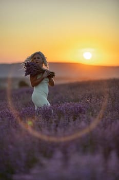 Blonde woman poses in lavender field at sunset. Happy woman in white dress holds lavender bouquet. Aromatherapy concept, lavender oil, photo session in lavender.