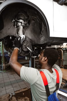 African male auto-mechanic repairing car brakes under the car in auto service close up