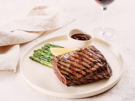 Grilled meat steak on plate with lemon and asparagus on white table background. Food and cuisine concept. Restaurant-style plating, with red wine glass on table