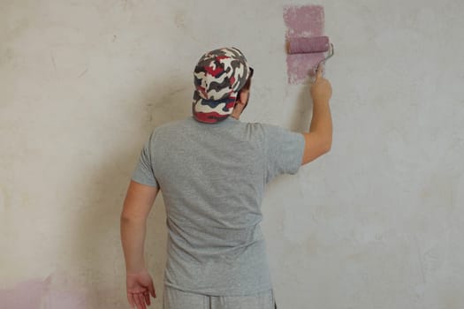 The rear view of a young adult man paints the wall with a roller. Repair in the house with their own hands.