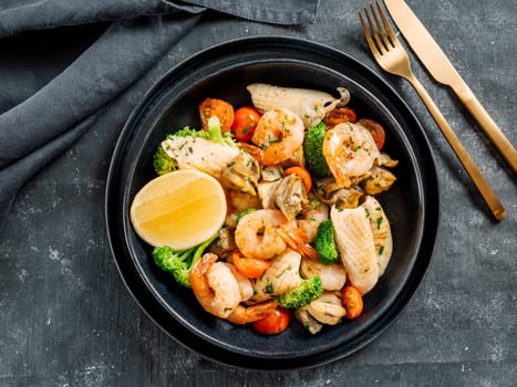 Mixed Seafood Contain Shrimps, Mussels, Calamari Squids and Fish with Broccoli, Cherry Tomato, Lemon on Black dish in restaurant-style platting. Seafood salad on dark background. Top view