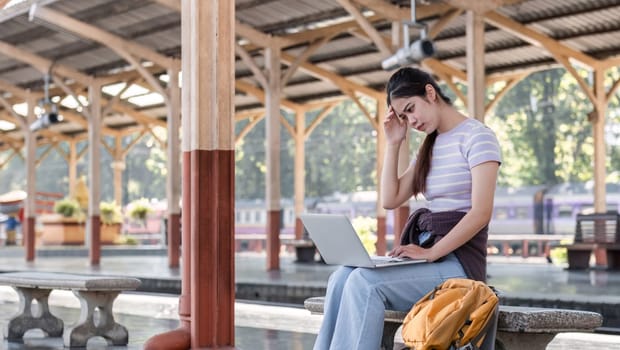 Young Asian woman in modern train station Female backpacker passenger sitting on a bench using a laptop while waiting for a train