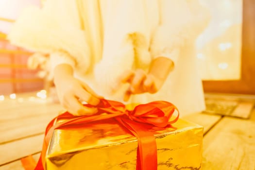 A woman in a white dress is holding a gold box with a red ribbon. She is wearing a crown on her head. The scene takes place in a room with a door and a window. The woman appears to be opening the gift box.