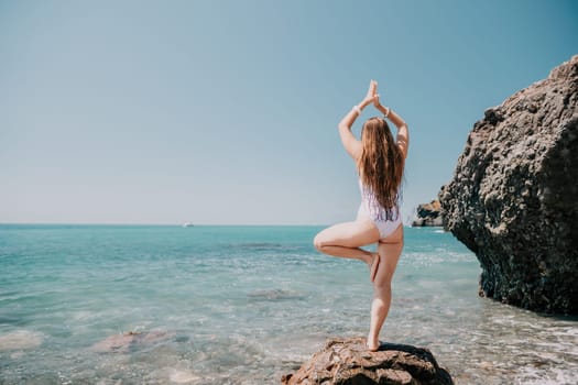 Woman sea yoga. Back view of free calm happy satisfied woman with long hair standing on top rock with yoga position against of sky by the sea. Healthy lifestyle outdoors in nature, fitness concept