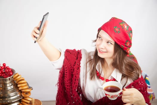 A fashionable modern girl in stylized folk clothes at table with a samovar, bagels and tea takes a selfie on the Orthodox holiday of Maslenitsa and Easter. Funny photo shoot for a young woman