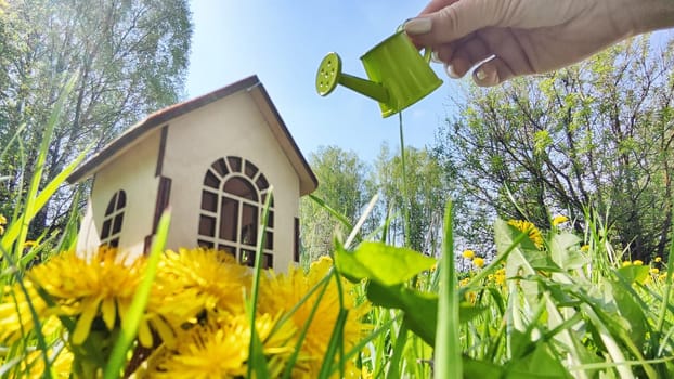 miniature toy house in grass, dandelion flowers and hand with watering can. natural background. symbol of Eco Friendly home, mortgage, construction, rental, property concept. Blurred, selective focus