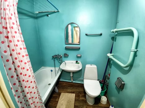 The interior of a small, modest bathroom with a bath and a toilet. Green walls, curtains with red dot. The concept of poverty and conciseness