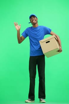 A male deliveryman, on a green background, full-length, with a box, waving his hand.