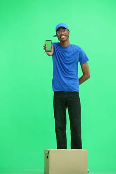 A male deliveryman, on a green background, full-length, with a box, shows a phone.