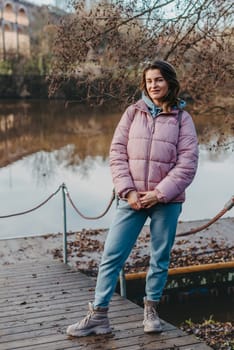 A young woman standing at the shore looking at the river in autumn sunny day. Street view, copy space for text, travel photo. Happy tourist woman on the bank of the river in autumn in warm clothes. Tourists enjoy their vacation, winter season. Romantic look and travel concept. A joyful mood in a Caucasian girl. Winter Wonderland: Enchanting Girl by the Riverside in Autumn.