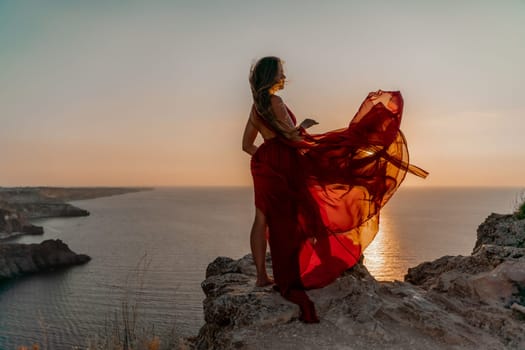 Woman sunset sea red dress, side view a happy beautiful sensual woman in a red long dress posing on a rock high above the sea on sunset