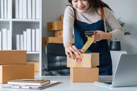 Young asian woman taping up a cardboard box in home office SME e-commerce business, relocation and new small business concept, SME concept.