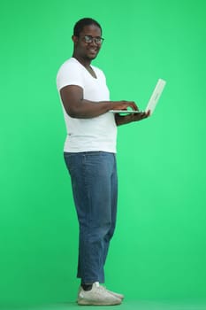 Full-length man in a white T-shirt typing on a laptop on a green background, in profile.