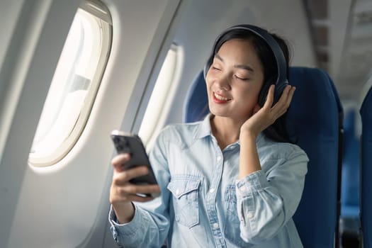 Asian woman traveler in airplane wearing headset listening music from mobile phone going on a trip vacation travel concept.