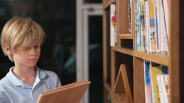 Young smart caucasian boy picking or choosing from book shelf and reading a book. Clever child learning, studying, open a books at library. Attractive kid turning page. Stack of books. Erudition.