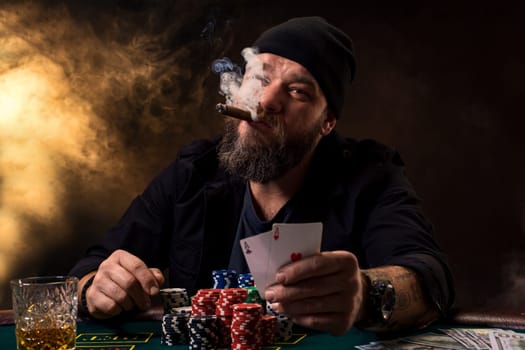 Bearded man with cigar and glass sitting at poker table in a casino. Gambling, playing cards and roulette. On the green poker table are cards, chips and money. The whole room is in smoke from cigars