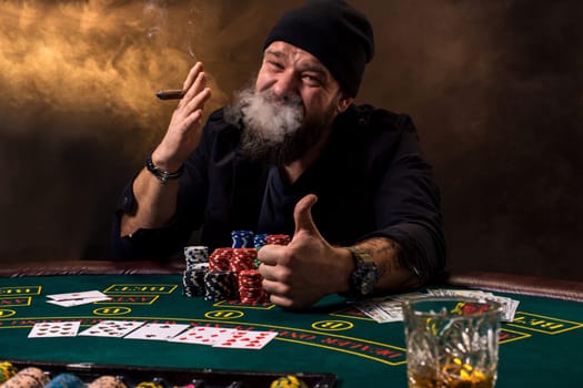 Bearded man with cigar and glass sitting at poker table in a casino. Gambling, playing cards and roulette. On the green poker table are cards, chips and money. The whole room is in smoke from cigars