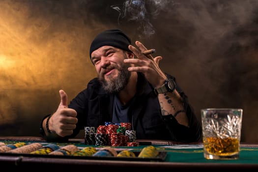 Bearded man drinking whisky while playing poker on the table with thick cigarette smoke. The concept of victory. The concept of gambling
