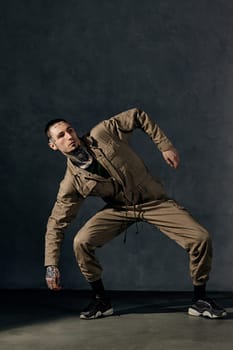 Young stylish guy with tattooed body and face, earrings, beard. Dressed in khaki overalls and black sneakers. He is dancing against gray studio background. Dancehall, hip-hop. Full length, copy space