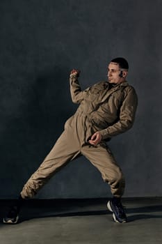 Young stylish performer with tattooed body and face, earrings, beard. Dressed in khaki overalls and black sneakers. He is dancing against gray background. Dancehall, hip-hop. Full length, copy space