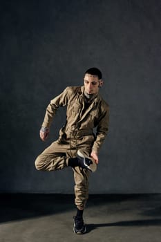 Young handsome man with tattooed body and face, earrings, beard. Dressed in khaki overalls and black sneakers. He is dancing against gray studio background. Dancehall, hip-hop. Full length, copy space