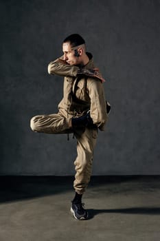 Young handsome male with tattooed body and face, earrings, beard. Dressed in khaki jumpsuit and black sneakers. He dancing against gray studio background. Dancehall, hip-hop. Full length, copy space