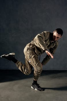 Young handsome fellow with tattooed body and face, earrings, beard. Dressed in khaki jumpsuit and black sneakers. He dancing against gray studio background. Dancehall, hip-hop. Full length, copy space