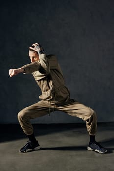 Young graceful man with tattooed body and face, earrings, beard. Dressed in khaki overalls and black sneakers. He is dancing against gray studio background. Dancehall, hip-hop. Full length, copy space