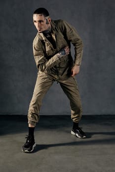 Young modern performer with tattooed body and face, earrings, beard. Dressed in khaki overalls and black sneakers. He is dancing against gray background. Dancehall, hip-hop. Full length, copy space