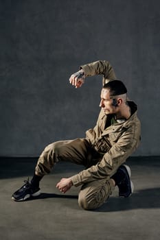 Young athletic man with tattooed body and face, earrings, beard. Dressed in khaki overalls and black sneakers. He is dancing against gray studio background. Dancehall, hip-hop. Full length, copy space