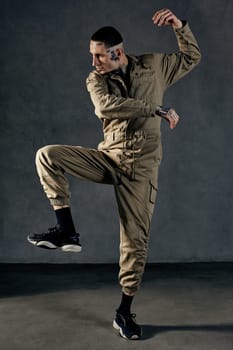 Young athletic male with tattooed body and face, earrings, beard. Dressed in khaki jumpsuit and black sneakers. He dancing against gray studio background. Dancehall, hip-hop. Full length, copy space