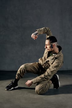 Young athletic guy with tattooed body and face, earrings, beard. Dressed in khaki overalls and black sneakers. He is dancing against gray studio background. Dancehall, hip-hop. Full length, copy space