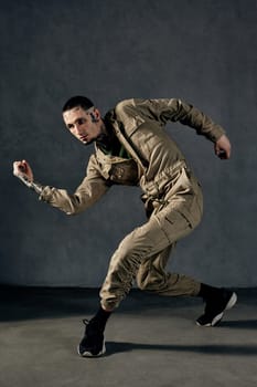 Young athletic performer with tattooed body and face, earrings, beard. Dressed in khaki overalls and black sneakers. He is dancing against gray background. Dancehall, hip-hop. Full length, copy space