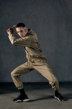 Good-looking male with tattooed body and face, earrings, beard. Dressed in khaki jumpsuit and black sneakers. He is dancing against gray studio background. Dancehall, hip-hop. Full length, copy space