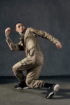 Good-looking guy with tattooed body and face, earrings, beard. Dressed in khaki overalls and black sneakers. He is dancing against gray studio background. Dancehall, hip-hop. Full length, copy space